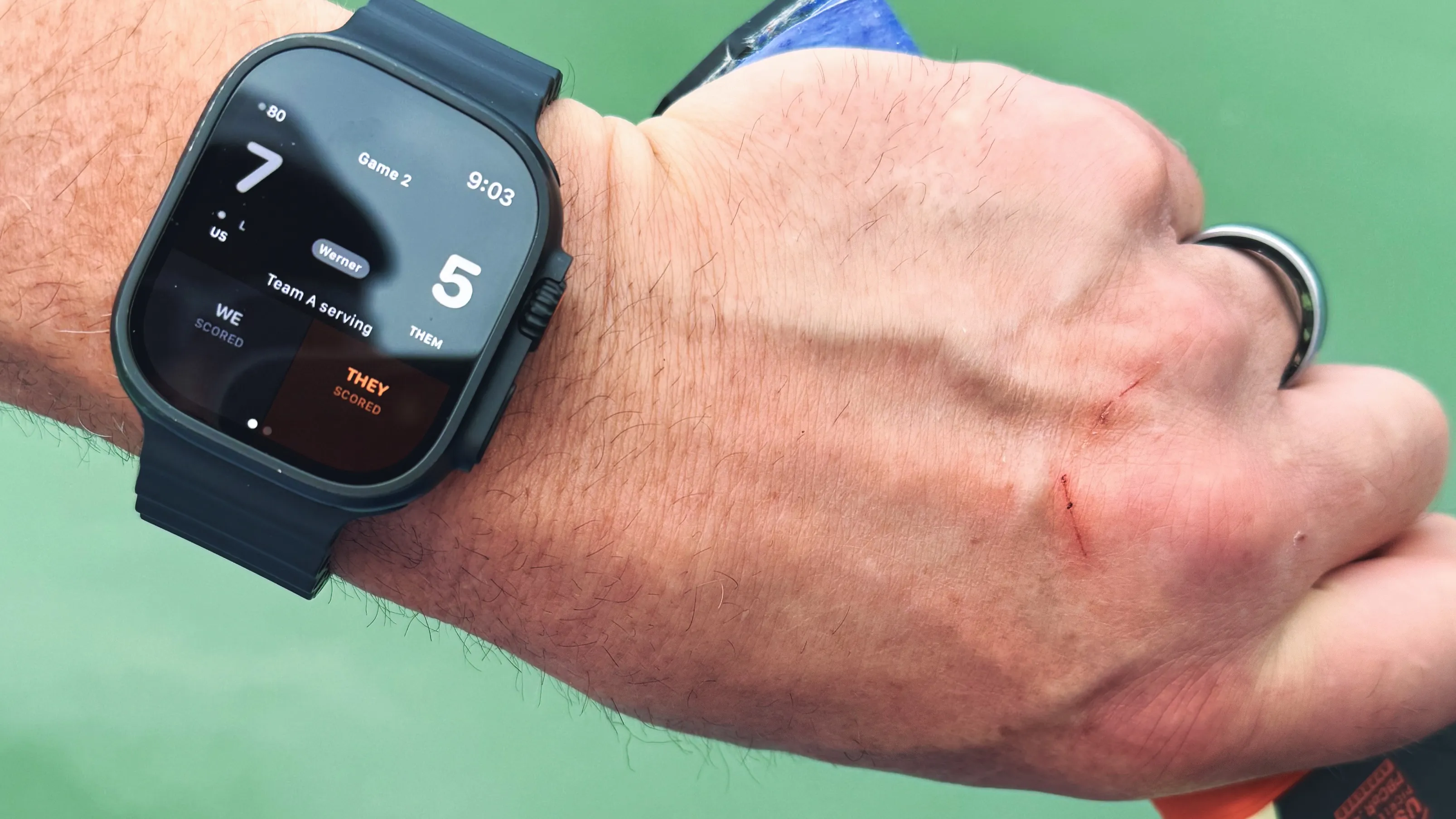 Apple Watch on wrist showing the CourtSauce Pickleball scoring UI mid-match, paddle visible in the background on a pickleball court