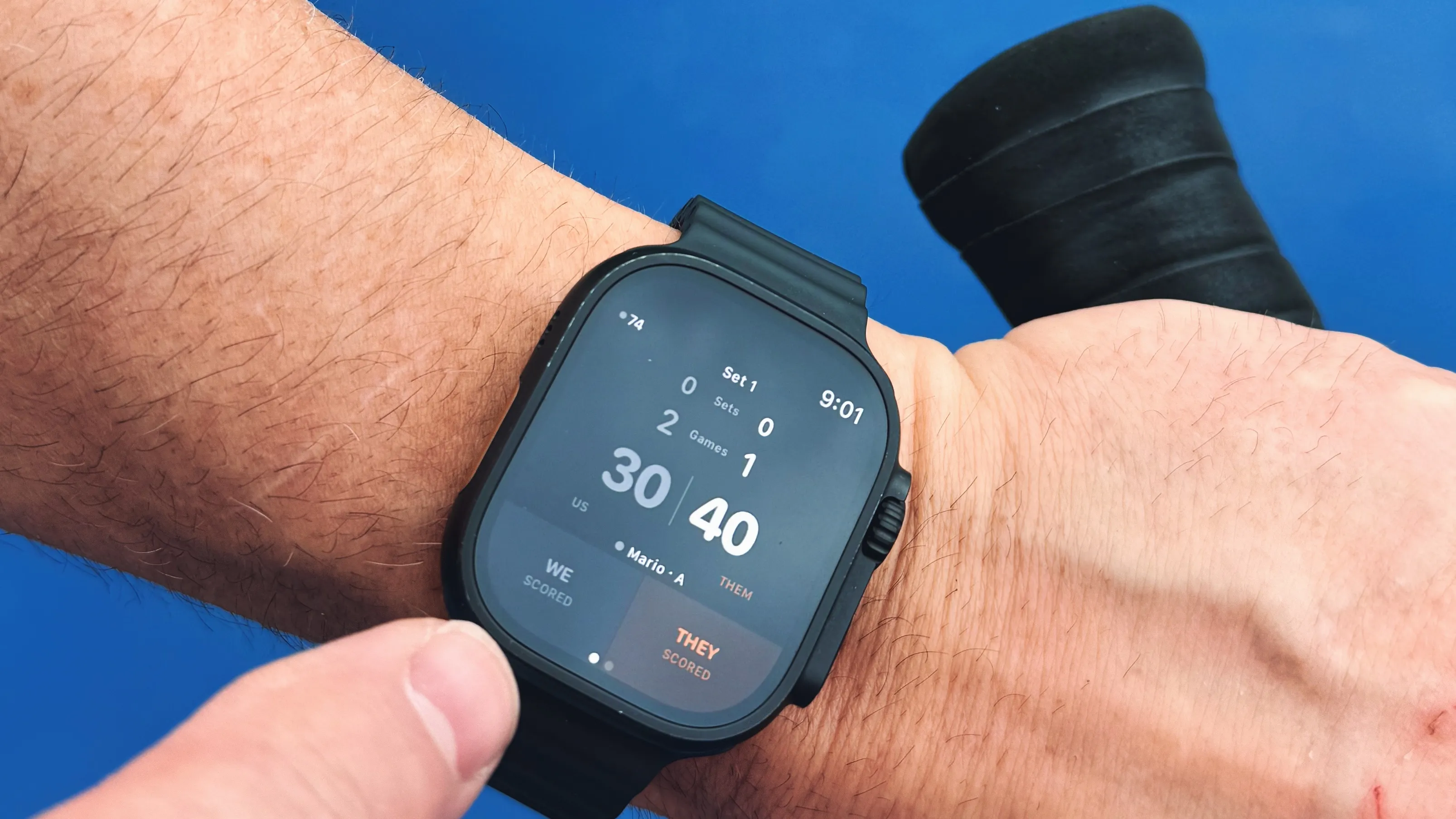 Apple Watch on wrist showing the CourtSauce Tennis scoring UI mid-match, tennis racquet visible in the background on a tennis court
